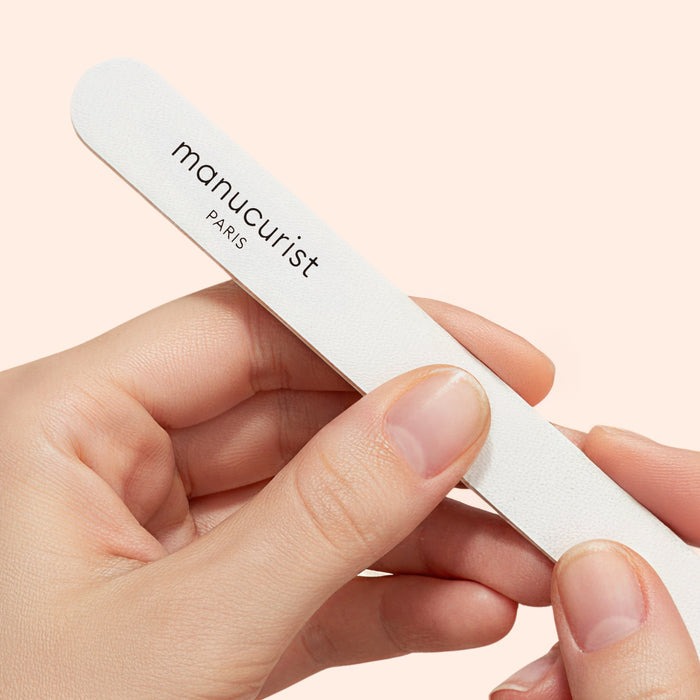 Close-up of a person shaping their sensitive nails with a Manucurist-us 180/240 emery board for fragile nails (pack of 5) against a light peach background.