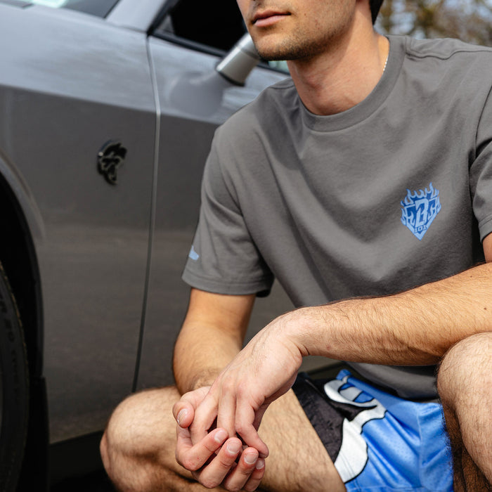 Man seated beside silver car, wearing gray Flame Thrower tee with blue BL101 flame diamond chest graphic and blue flame shorts.