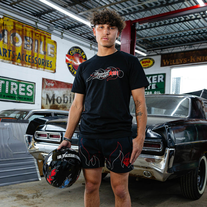Young man standing in vintage garage holding racing helmet, wearing black Flames Stitched tee with BL101 flame graphic and black flame shorts, classic cars in background.