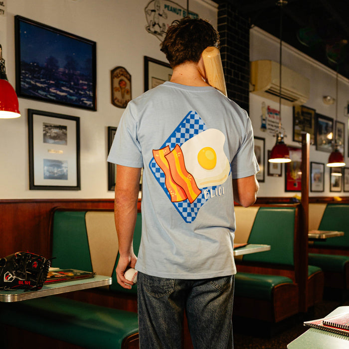 Person wearing a light blue t-shirt with a graphic design of an egg and bacon and BL101 logo in a diner.