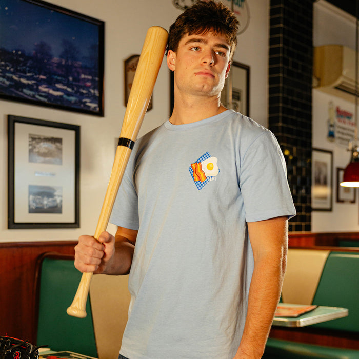Man wearing a light blue t-shirt with bacon and eggs on it, holding a baseball bat in a diner.