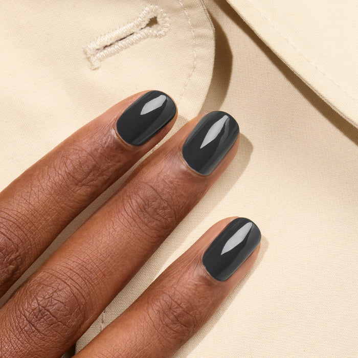 A close-up of three fingers with glossy, dark grey Manucurist Anthracite Green Flash gel polish resting on beige fabric with visible stitching and a buttonhole.