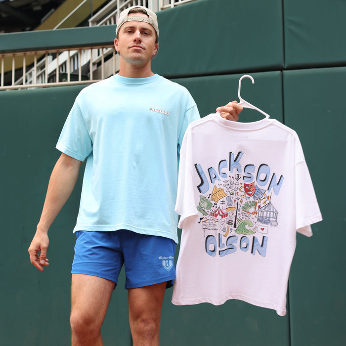 Adult male model wearing the light blue Jackson Olson x BL101 BSBL Era relaxed tee while holding the white Hometown Relaxed Tee on a hanger