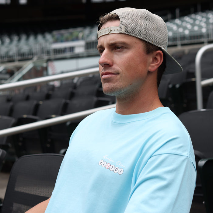 Adult male model seated in stadium seating wearing the light blue Jackson Olson x BL101 BSBL Era relaxed tee with chest logo