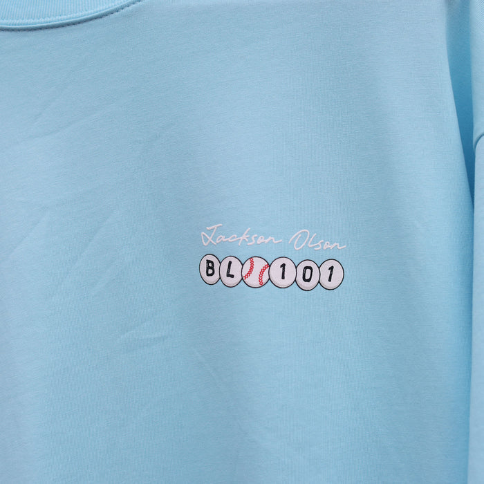 Close-up of the chest graphic on the light blue BSBL Era tee, showing the "Jackson Olson" script above the BL101 baseball bead logo