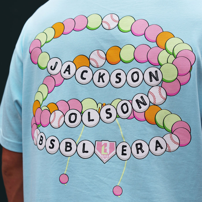 Close-up of the back graphic on the light blue BSBL Era tee, featuring a colorful friendship bracelet design with "Jackson Olson BSBL Era" lettering and baseball bead details