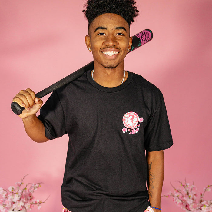 Male model wearing a black BL101 cherry blossom tee and holding a baseball bat over his shoulder against a pink background, front view