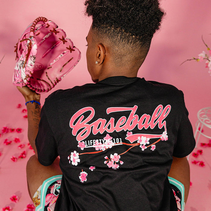Male model seated against a pink background holding a pink floral baseball glove, showing the back of a black BL101 tee with pink "Baseball" script and cherry blossom graphic