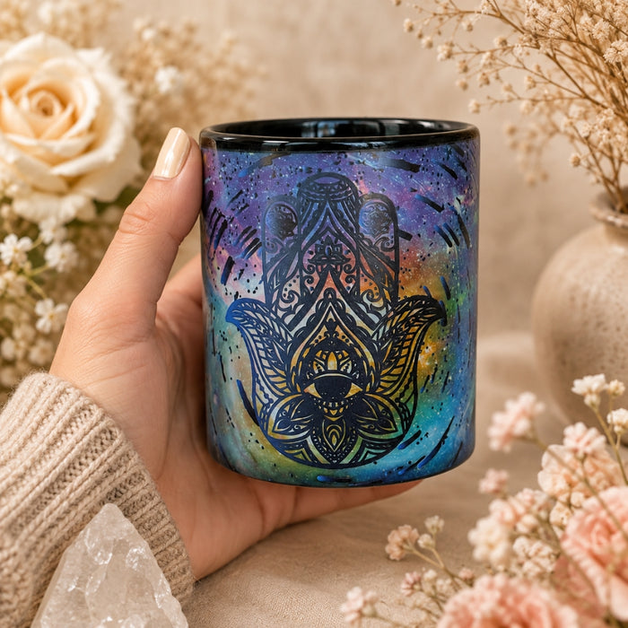 A hand holding a ceramic mug decorated with a black Hamsa hand design over a blue, purple, and teal galaxy watercolor background, surrounded by dried flowers and a neutral beige setting.