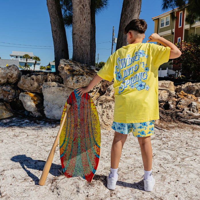 Person in yellow shirt with 'Swing into Spring' text and floral shorts holding a colorful board on a beach.