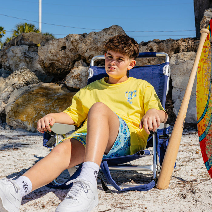 Child sitting on a beach chair wearing yellow shirt with floral BL101 logo by rocks with a surfboard nearby