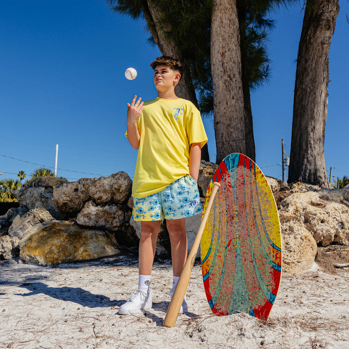 Person in yellow BL101 shirt and floral shorts holding a baseball and bat on a beach with a colorful surfboard.