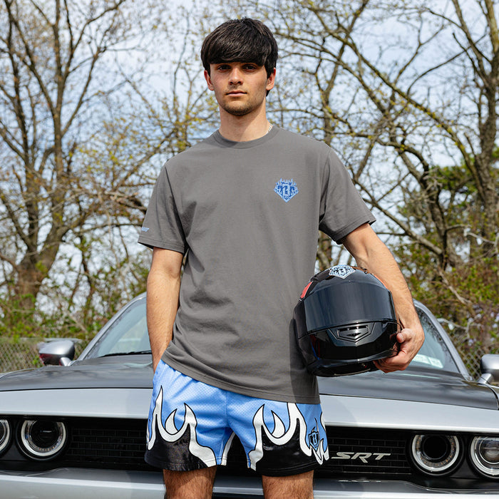 Young man standing in front of silver Dodge SRT Challenger holding racing helmet, wearing gray Flame Thrower tee with blue BL101 chest graphic and blue flame shorts.