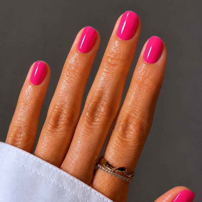 A hand with neatly manicured nails painted bright pink using LONDONTOWN's Flash Dry Top Coat, wearing two gold rings, set against a plain gray background.