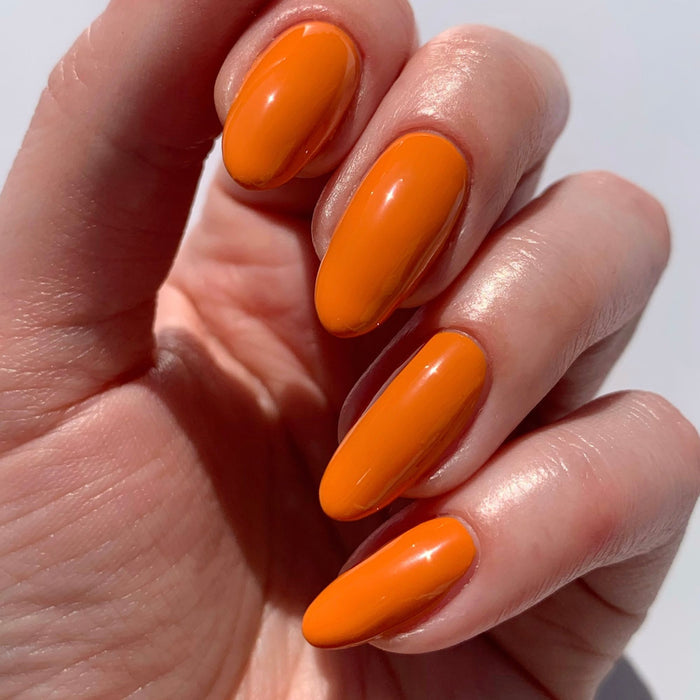 A hand with long, almond-shaped nails painted in a glossy bright orange, finished with LONDONTOWN's Flash Dry Top Coat for a fast-dry shine.