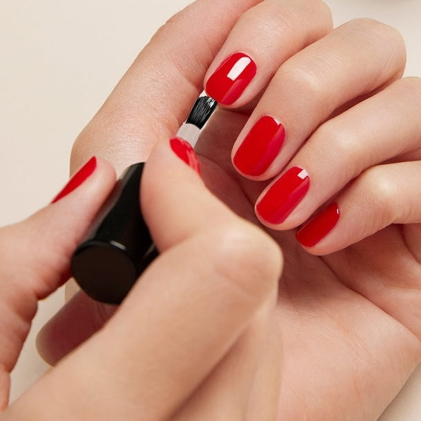 A person applies Manucurist Green Sunshine Top Coat to their fingernails, holding a brush in one hand and painting the already partially polished nails for a glossy, plant-based gel effect.