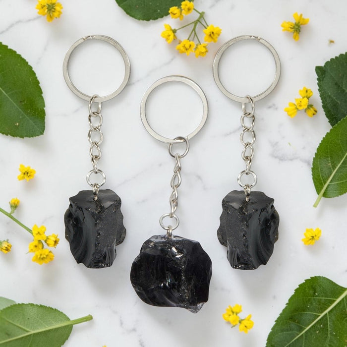 Three black Obsidian keychains on a marble surface with green leaves and yellow flowers.