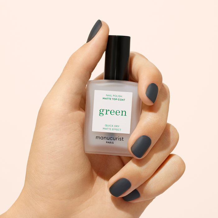 A hand with short, matte dark gray nails holds a bottle of Manucurist Matte Top Coat, a plant-based polish ideal for eco-friendly manicures, against a light beige background.