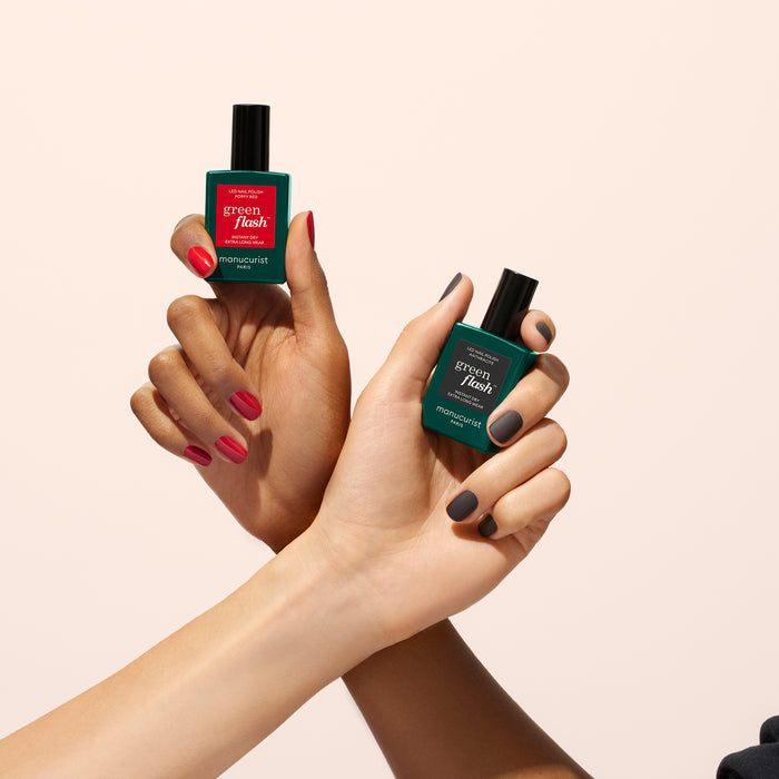 Two hands with manicured nails hold green bottles of Manucurist Matte Top Coat; one hand has red nails, the other dark gray, both showcasing a plant-based formula against a plain light background.