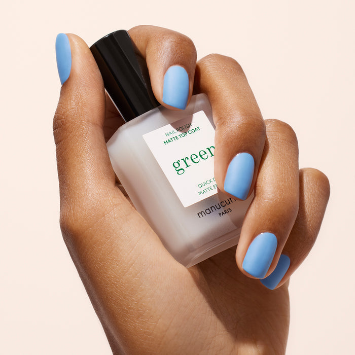 A hand with light blue nails holds a frosted bottle of Manucurist Matte Top Coat nail polish with a black cap, perfect for an eco-friendly manicure, against a light background.