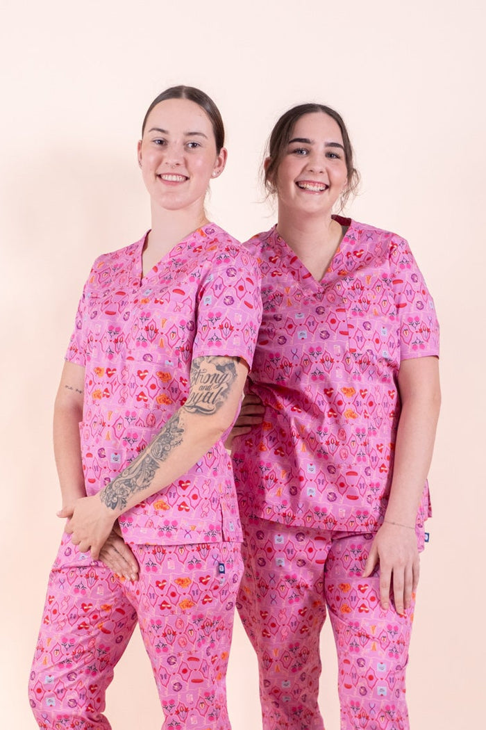 Two women modeling matching pink medical scrubs with a small floral pattern, standing against a solid pale peach studio background.