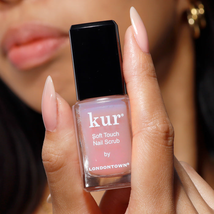 A person with manicured nails holds a bottle of LONDONTOWN's Soft Touch Nail Scrub, an ideal gentle exfoliator for refining nails and cuticles.
