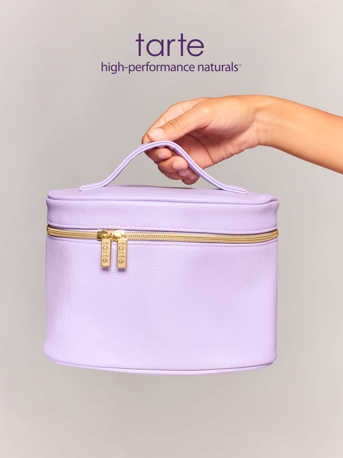 A lavender makeup bag with a handle is held by a hand, showcasing a zippered closure with two gold zippers labeled Tarte, against a plain, light gray background.