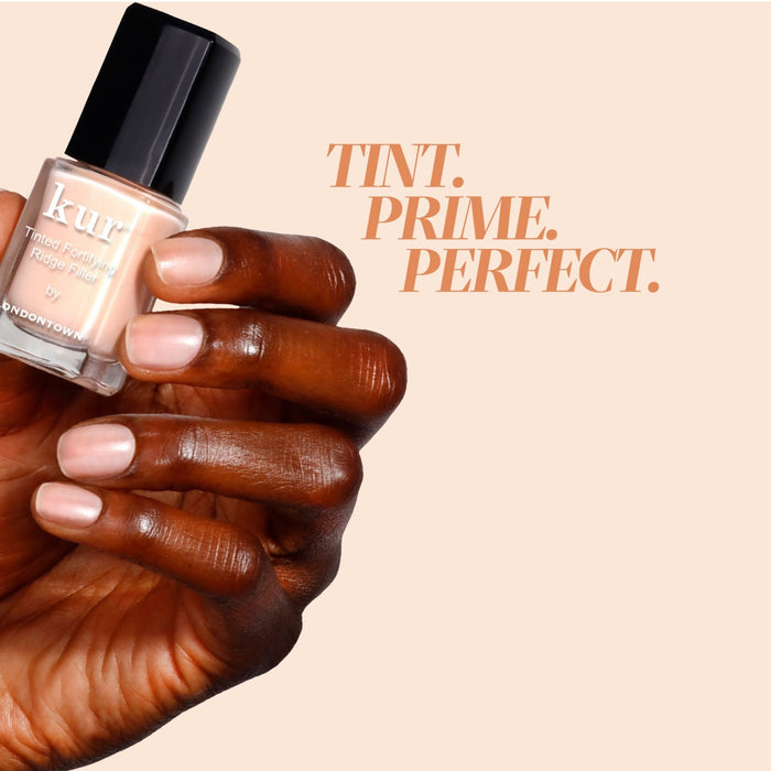 A hand holds LONDONTOWN’s Tinted Fortifying Ridge Filler in Apricot Beige against a beige background with “Tint. Prime. Perfect.” in bold, showing how it smooths ridges for an even finish and a subtle hint of tint.