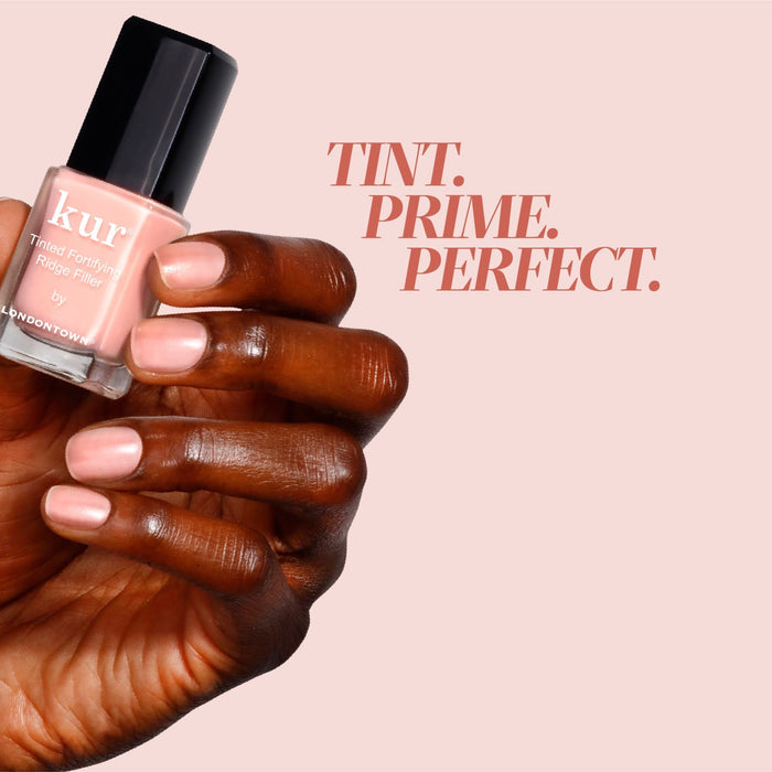 A hand holding LONDONTOWN’s Tinted Fortifying Ridge Filler in Petal Pink with manicured nails; text reads "TINT. PRIME. PERFECT." on a pale pink background, highlighting smooth ridges and a hint of tint for flawless nails.